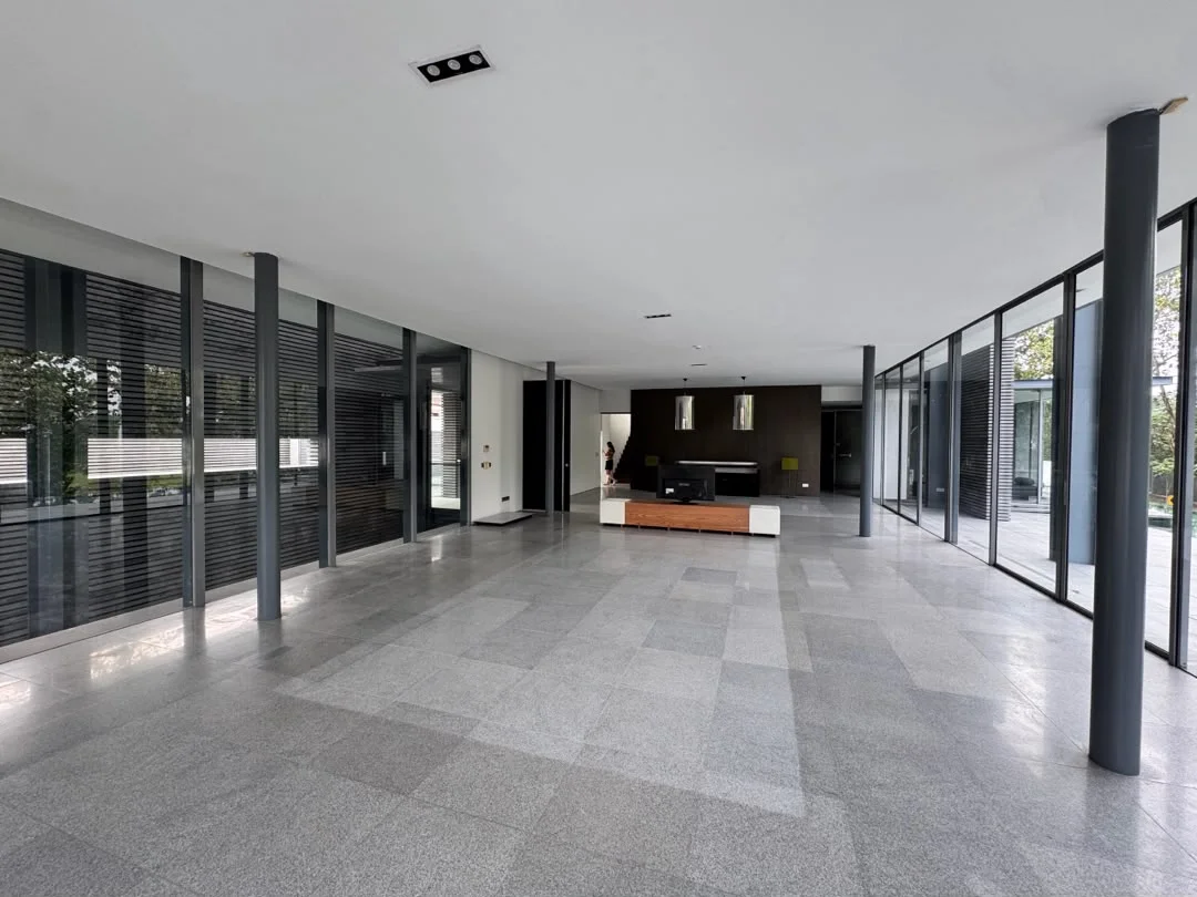Room features: large open space, grey polished stone flooring, floor-to-ceiling windows, black accent wall, minimalist design, exposed structural columns, natural light flooding in, sliding glass doors, view of outdoor greenery; style: modern; view: garden.